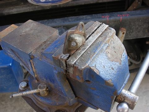 1987 Ford Bronco - Close-up of a chipped blue bench vise clamped on a shop bench, with a small metal part gripped in its serrated steel jaws and a blue Ford o…