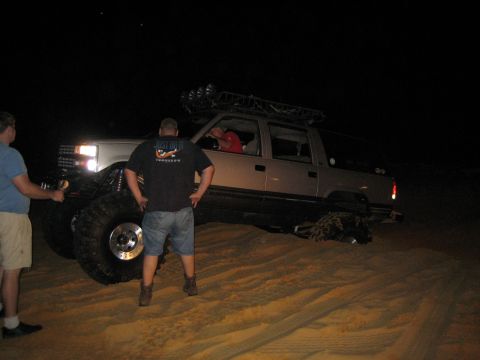 1989 Ford Bronco - A modified truck with large tires is parked on sand dunes at night, with people standing nearby.