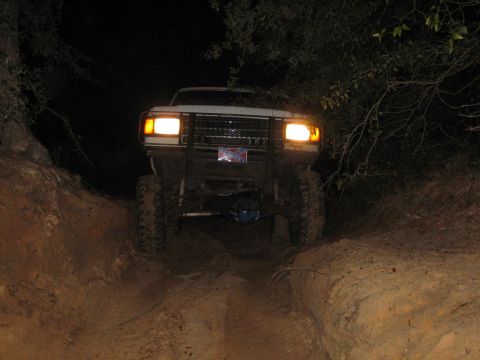 1989 Ford Bronco - A white Ford Bronco navigates a dirt trail in the dark, its headlights illuminating the path ahead.