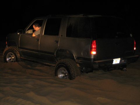 1989 Ford Bronco - A dark SUV is stuck in the sand, seen from the 3/4 front angle. The vehicle appears to be from the mid-1990s.