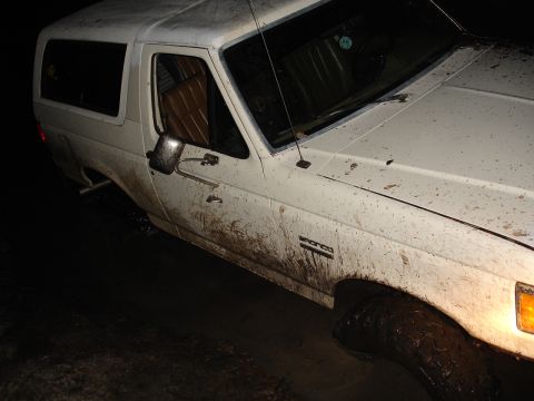 1989 Ford Bronco - A white 1989 Ford Bronco is stuck in mud, with the front 3/4 view highlighting the mud-covered exterior and tires.
