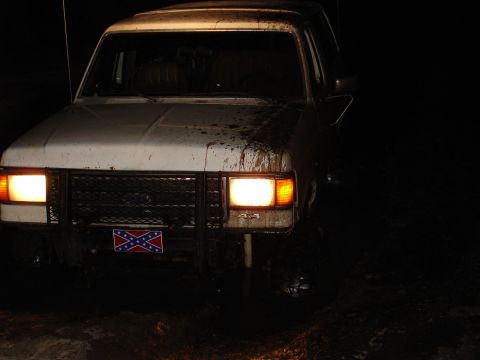 1989 Ford Bronco - Mud-covered 1989 Ford Bronco with headlights illuminated, seen at night.