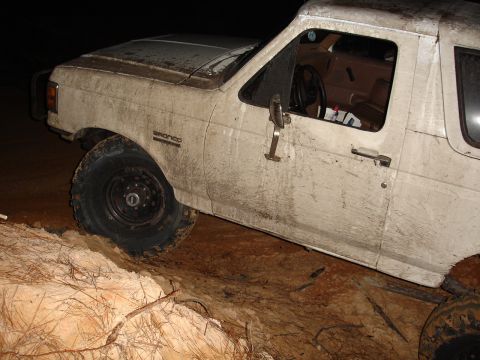 1989 Ford Bronco - A white 1989 Ford Bronco is shown partially stuck in mud and dirt, with its tires digging into the terrain.