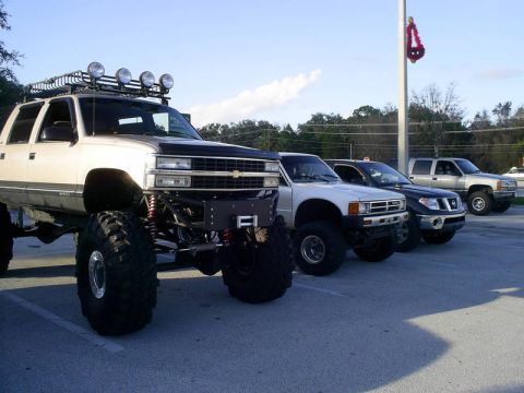 1989 Ford Bronco - Several modified trucks are parked in a lot, including a heavily lifted beige Chevrolet Tahoe in the foreground.