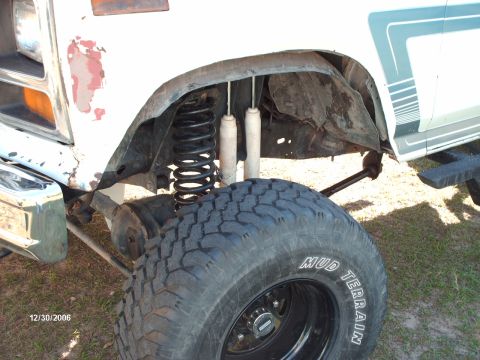 1982 Ford F-100 - Close-up of the front driver-side suspension and wheel well of a white Ford pickup with faded blue side stripes, showing a black coil sprin…
