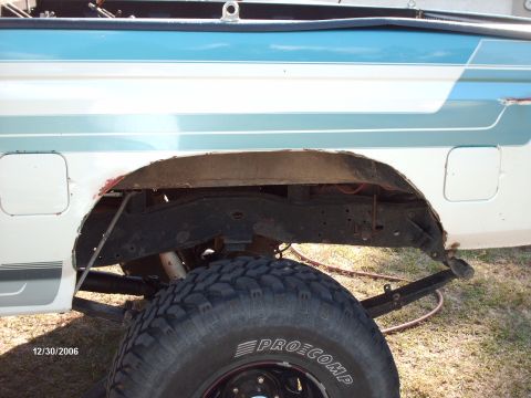 1982 Ford F-100 - Close-up of the rear wheel well of a two-tone teal and white Ford pickup with the wheel/tire removed, exposing the bare metal fender, frame…