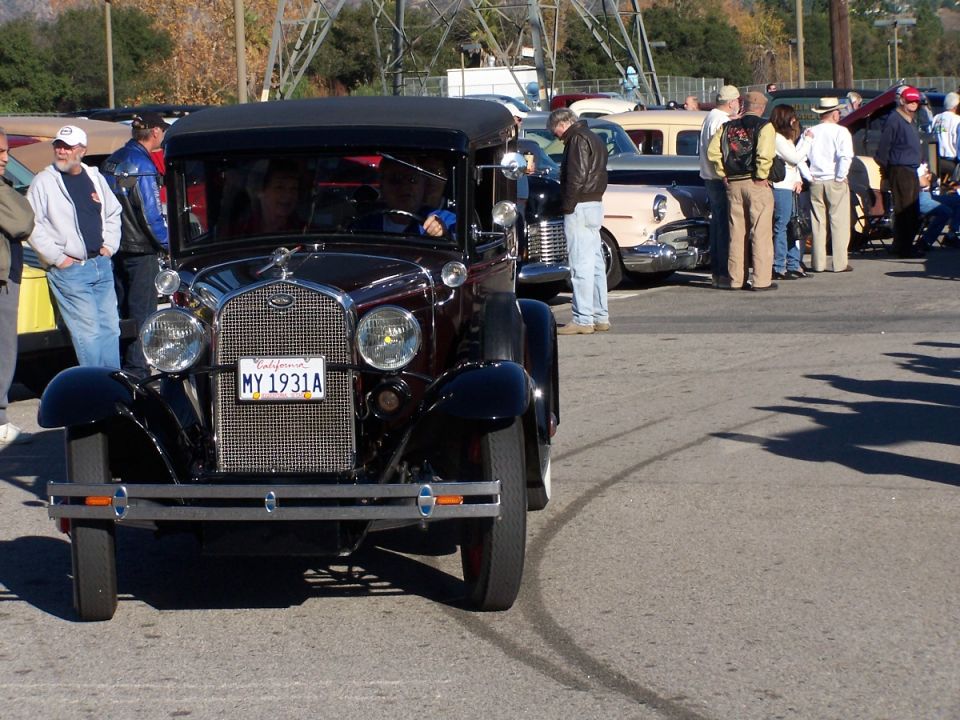 Front view of a dark-colored vintage Ford sedan with a "MY 1931A" license plate. Other classic cars and people are visible in the background