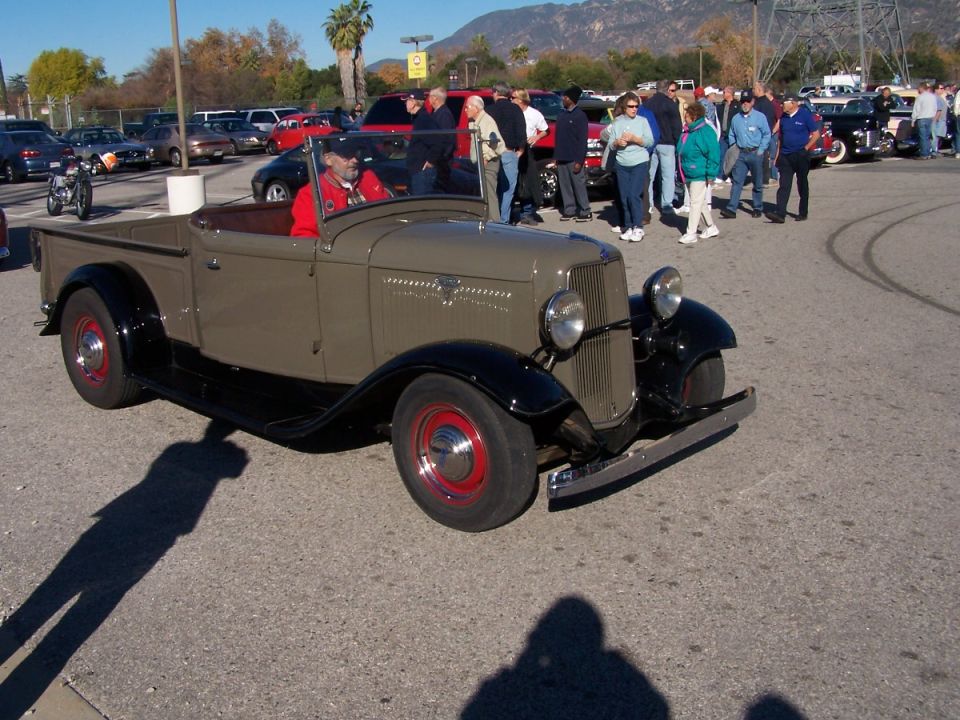 Tan and black 1930s Ford pickup truck with red wheels, driver visible, parked outdoors