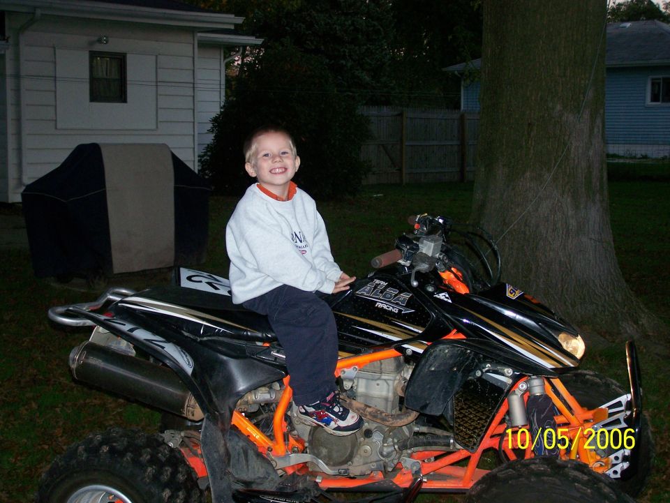 Young boy sitting on a black and orange sport ATV with Alba Racing graphics in a backyard