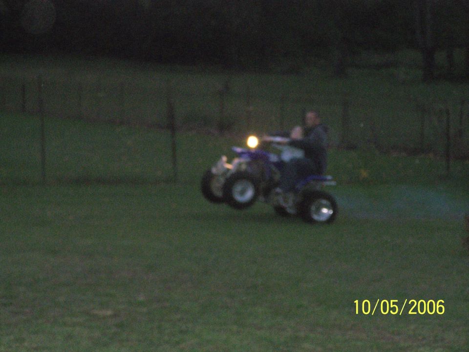 Two riders on a blue ATV with headlight on in a grassy field at dusk, date stamp 10/05/2006