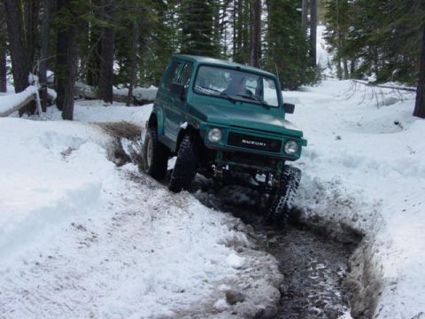 1986 Ford Bronco - A green Suzuki Samurai drives through a snowy and muddy path surrounded by trees.