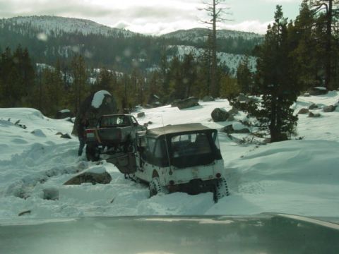 1986 Ford Bronco - Two vehicles are stuck in the snow on a wooded mountainside, with one appearing to be a Jeep-like vehicle and the other a pickup truck.