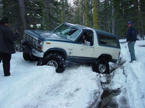 1986 Ford Bronco - A two-tone Ford Bronco navigates a snowy trail, with one wheel buried in a deep snowbank.