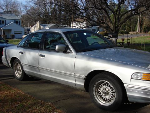 2003 Ford Crown Victoria - A silver 2003 Ford Crown Victoria is parked on a paved surface in front of a residential house.