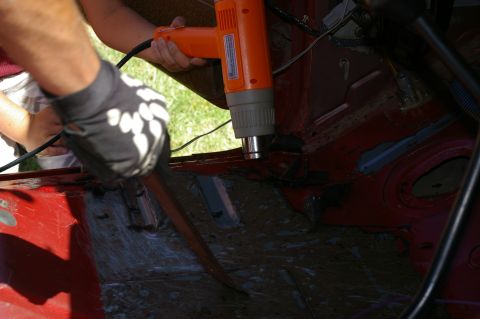 1986 Ford Bronco - A person uses a heat gun and scraper to remove old sound deadening from the floor of a red vehicle's interior.
