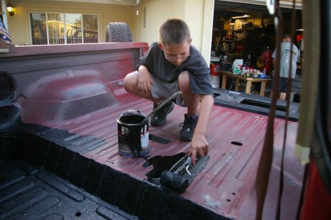 1986 Ford Bronco - A boy applies black coating to the bed of a red pickup truck.