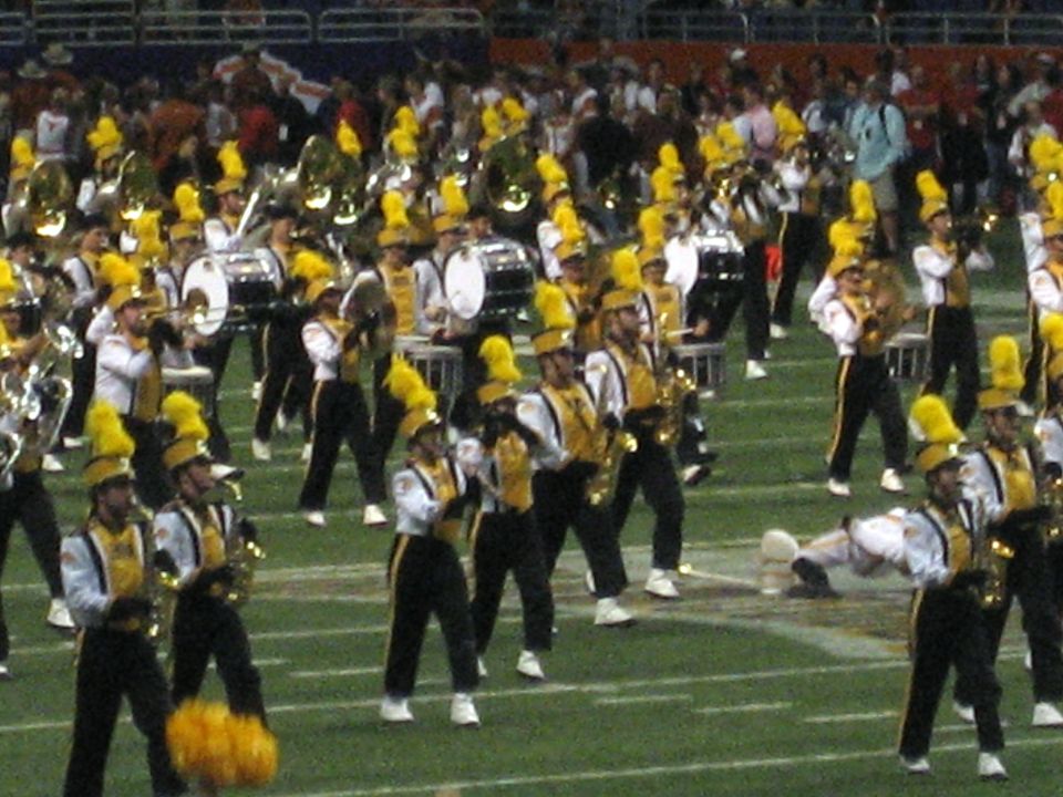 Marching band members in yellow and black uniforms with yellow hats perform on a football field