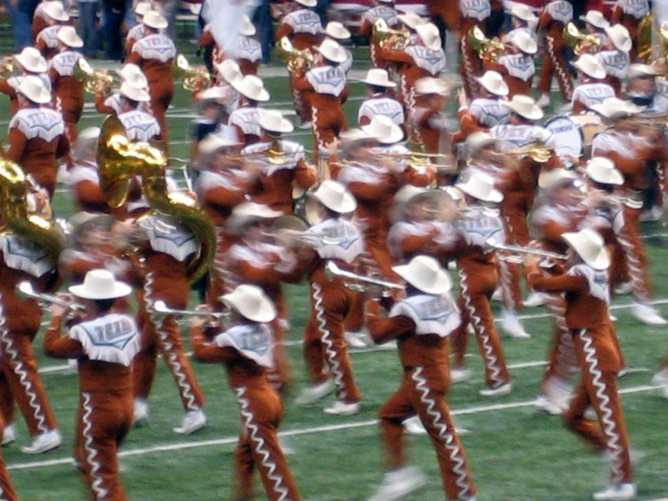 Marching band members in cowboy hats and brown uniforms play instruments on a football field