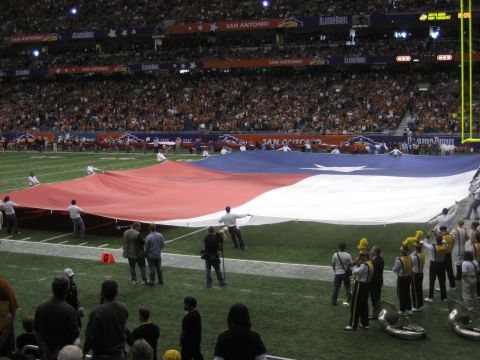 1986 Ford Bronco - People hold a large Texas flag on a football field during an event, with a crowd and marching band present.