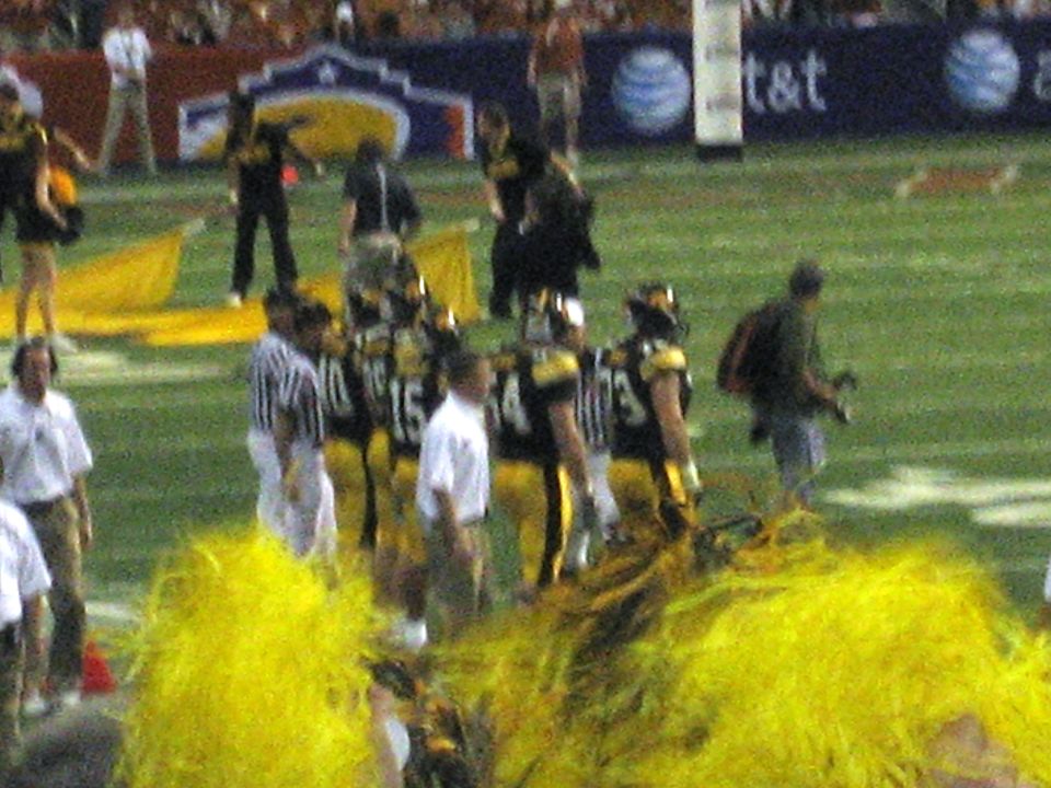 Football players in black and yellow uniforms on a green field with yellow pom-poms in the foreground