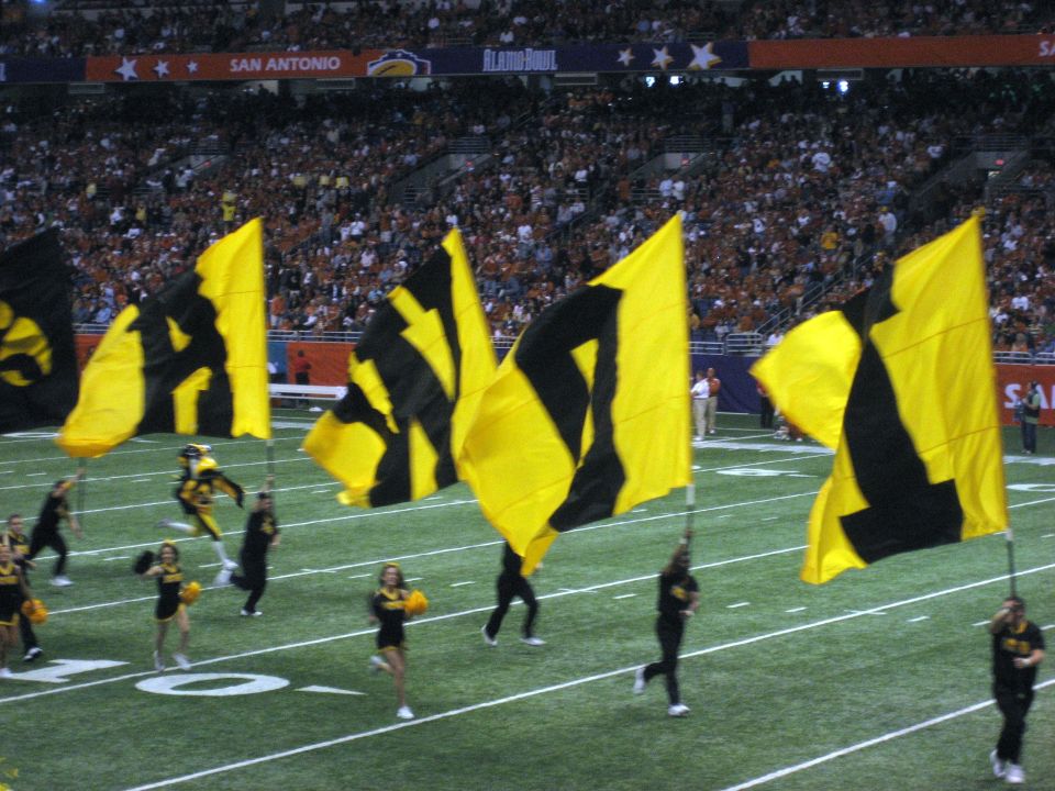 Football field with cheerleaders and large yellow and black flags, San Antonio Alamo Bowl banner in background