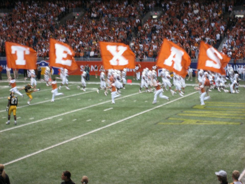 Football players run onto a field, holding large orange flags spelling 'TEXAS'. Spectators fill the stands