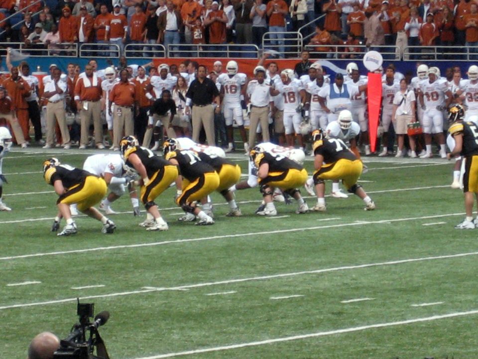 Football players in black and yellow uniforms on a green field, with spectators in the background