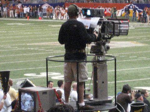 1986 Ford Bronco - A camera operator stands on a platform on a football field, ready to film the game.