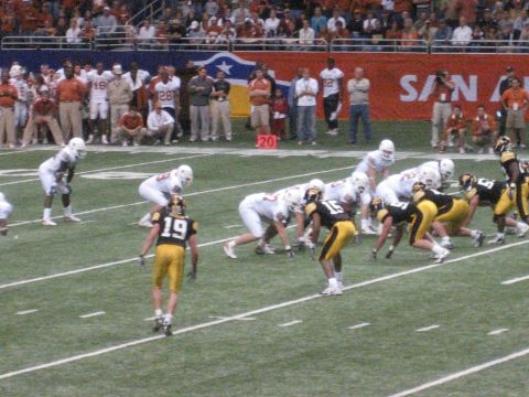 1986 Ford Bronco - Players in white and yellow uniforms face off on a football field with spectators in the background.