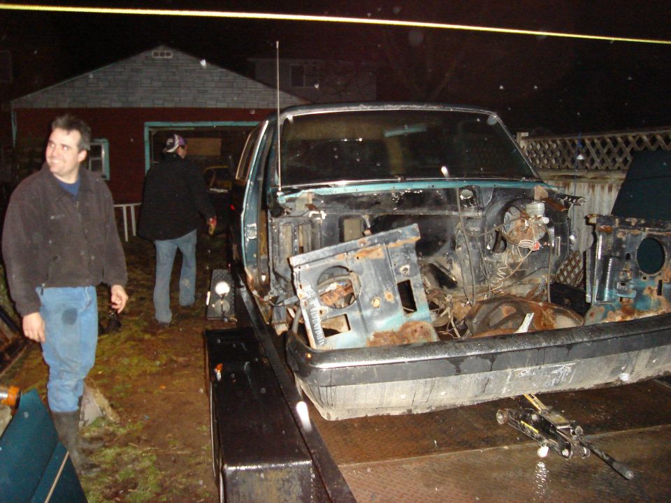 Teal car chassis on a trailer, front end dismantled, showing rust and exposed engine bay