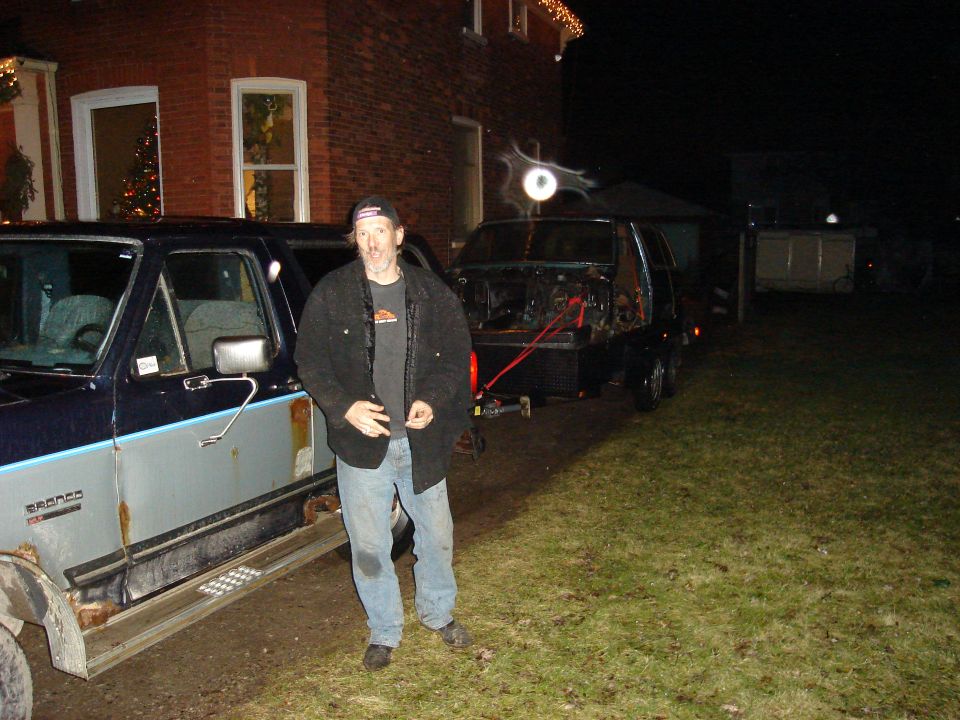 Man standing next to a rusty 1990 Ford Bronco, with another vehicle on a trailer behind it