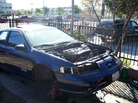 1992 Ford Taurus - A blue sedan, possibly a Ford Taurus, is parked with its hood open and missing a headlight, supported by jack stands.