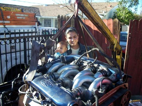 1992 Ford Taurus - A Ford engine is being lifted by a crane, with a woman and a baby watching.