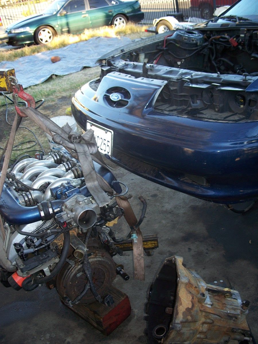 Engine removed from dark blue car, transmission nearby. Green car in background