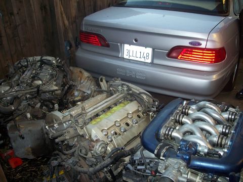 1992 Ford Taurus - A silver 1992 Ford Taurus is parked behind a collection of engines, suggesting a motor swap or parts storage.