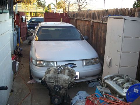 1992 Ford Taurus - A white 1992 Ford Taurus is parked in a narrow driveway, with engine components laid out in front and another vehicle visible behind it.