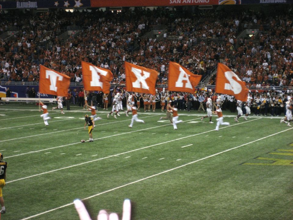 Football field with orange flags spelling TEXAS, players running, and a crowd in the stands