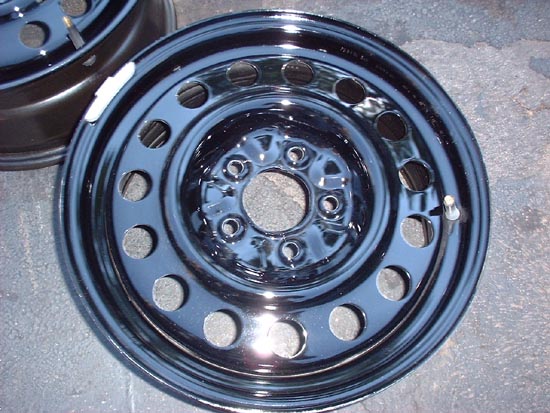 Close-up of a shiny black steel wheel with multiple lug nut holes. Another wheel is partially visible in the background