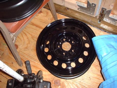 1997 Ford Crown Victoria - A black steel wheel is shown on a wooden surface, with a car jack and a blue cover nearby.