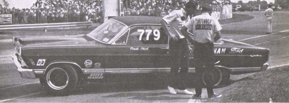 1960s Ford drag car number 779 in profile with two men at a race track, black and white.