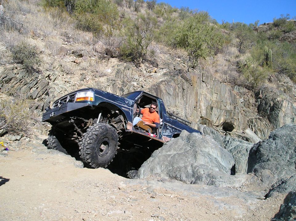 Dark blue truck with lifted suspension and large tires navigating rocky terrain. Driver in orange shirt visible