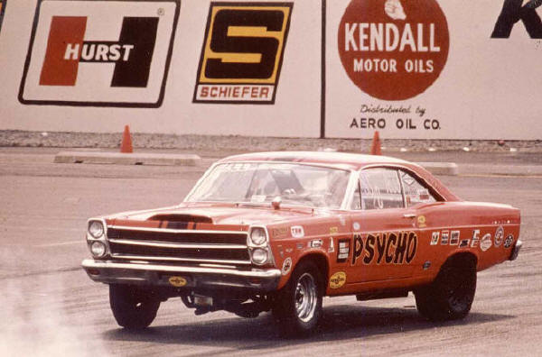 Orange vintage Ford drag coupe lettered 'PSYCHO' on a track with Hurst and Kendall signs behind.