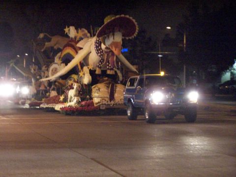 1990 Ford Bronco - A blue SUV drives past a parade float at night, its headlights illuminating the road ahead.