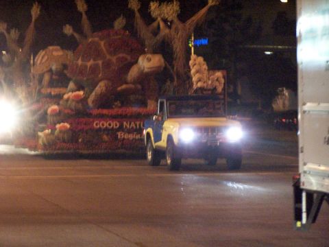 1990 Ford Bronco - A yellow Ford Bronco with a blue top drives past a float at night.