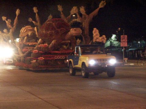 1990 Ford Bronco - A two-tone Ford Bronco drives past a float decorated with turtles and trees at night.