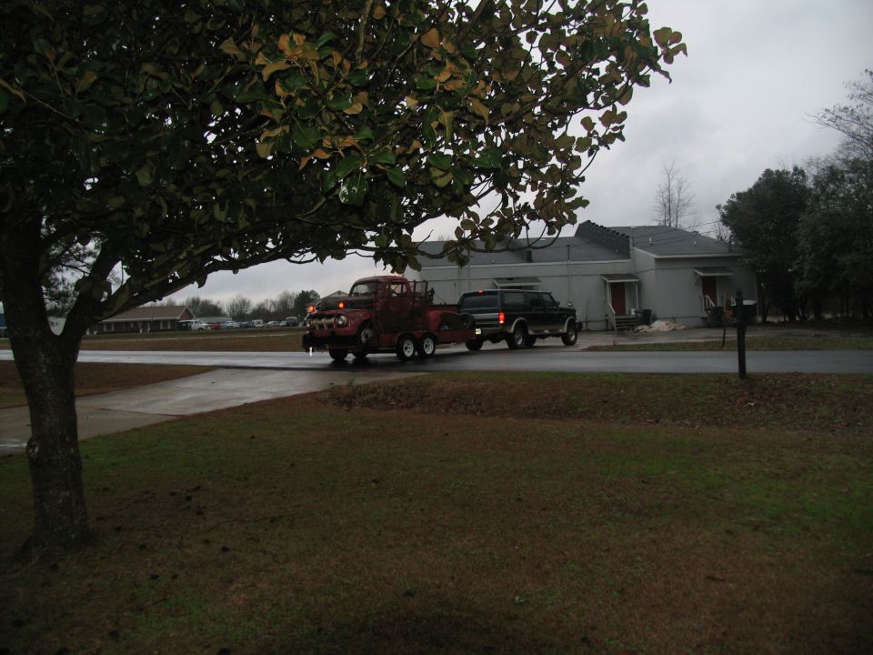 Weathered red 1950s Ford pickup on a tandem-axle trailer towed down a wet street