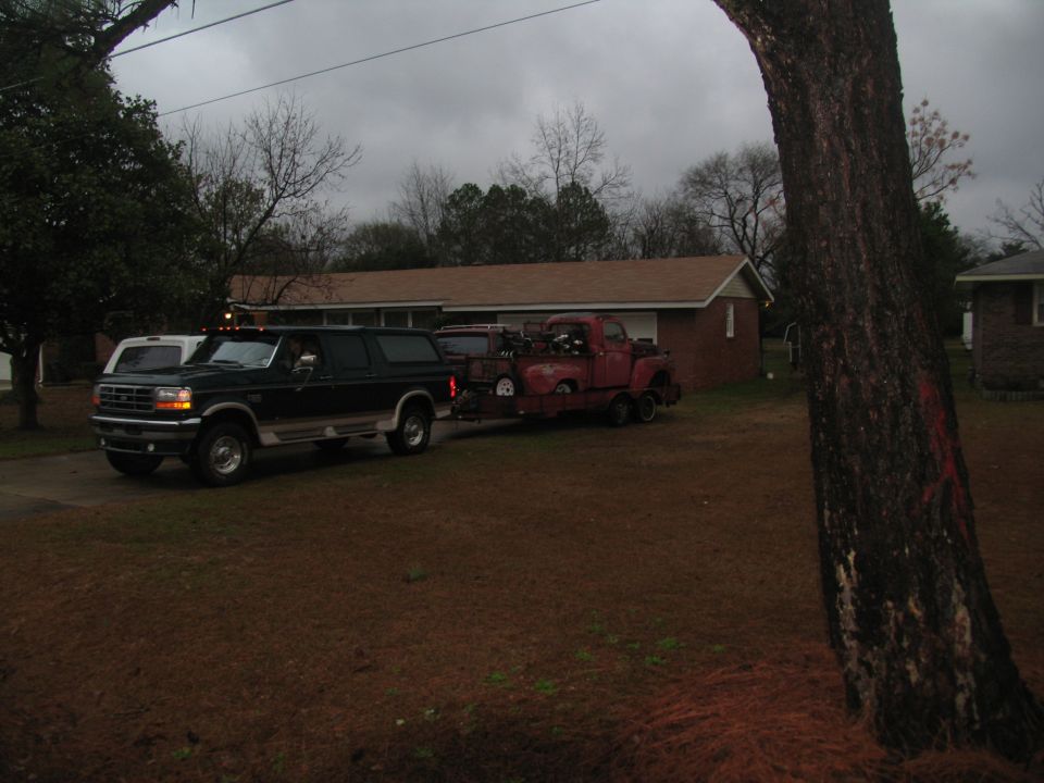Red pickup truck with dual rear wheels being towed by a dark SUV. The truck appears to be from the early 1950s