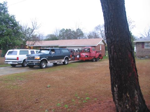 1952 Ford Pickup - A green SUV tows a red pickup truck on a trailer, with a white SUV parked in the driveway.