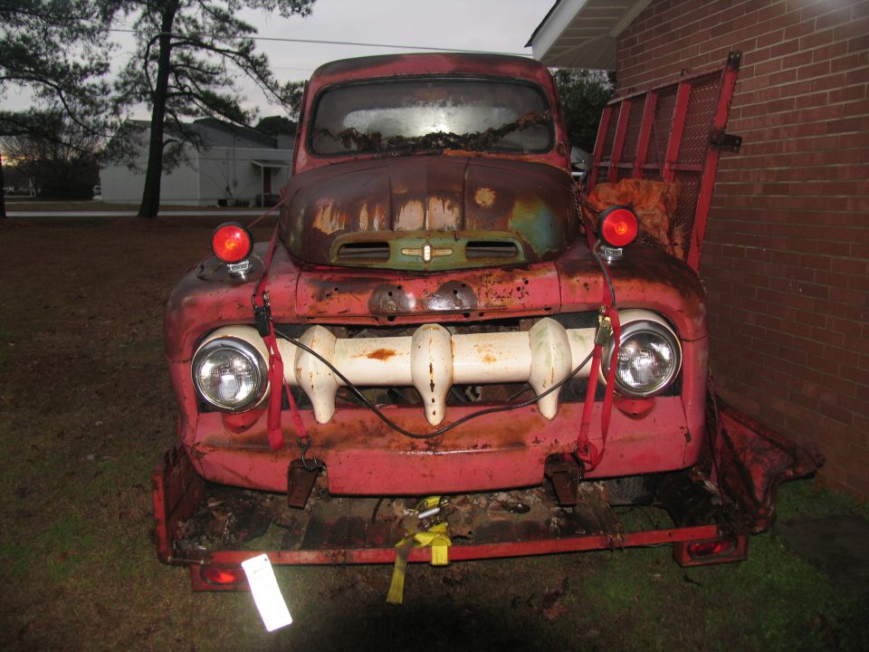 Rusty red 1950s Ford pickup front end strapped to a red trailer at dusk