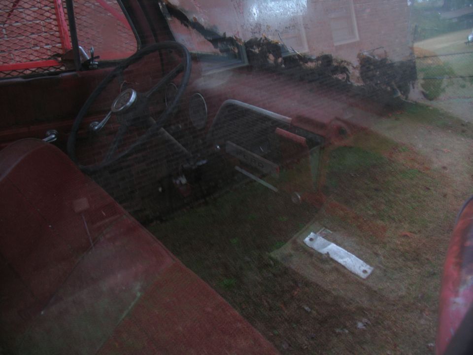 Interior of an old truck, showing the steering wheel, dashboard, and worn seats. The floor is covered in debris